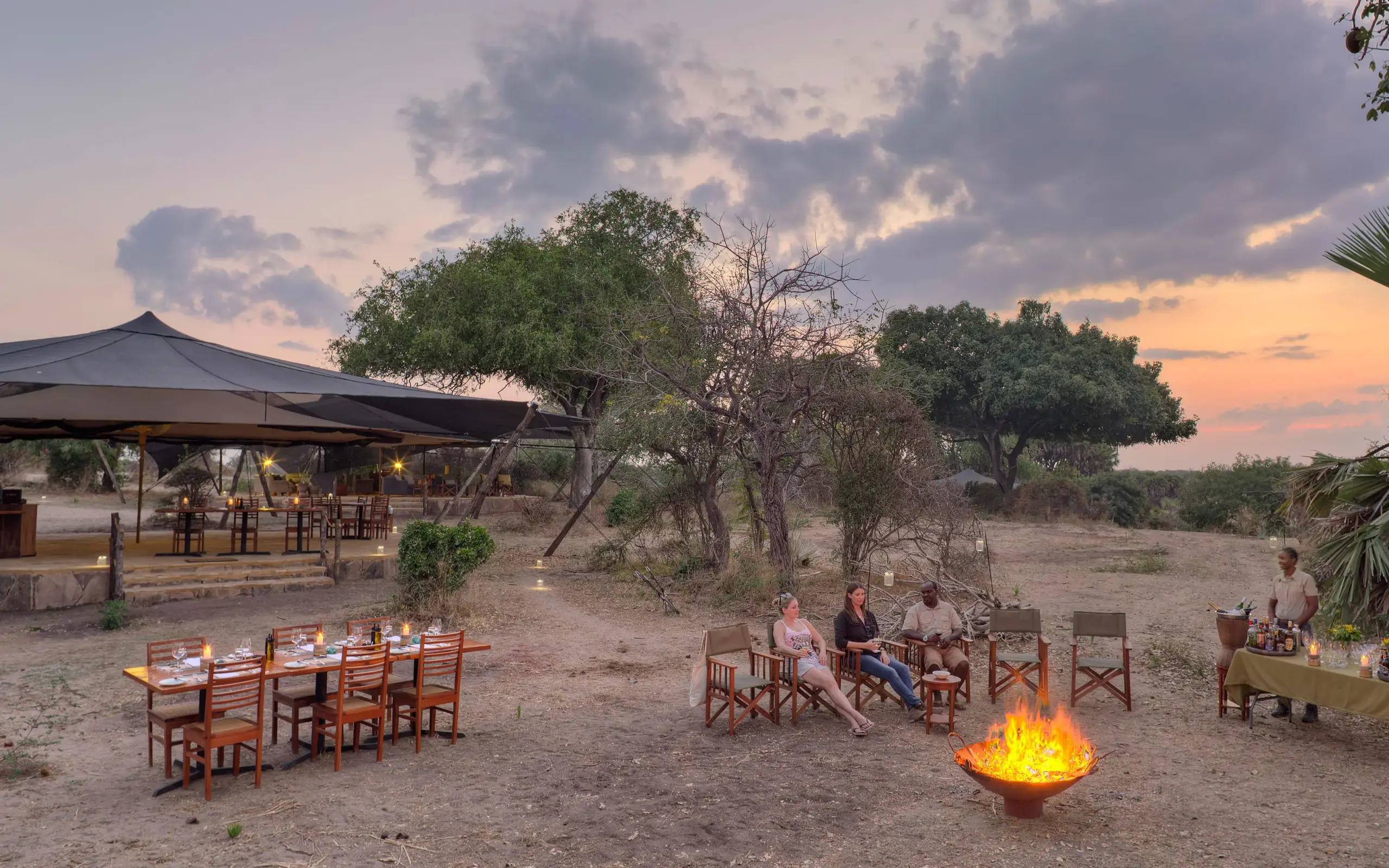 Safari camp outdoor dining area at sunset with campfire, Roho ya Selous, Nyerere National Park, Tanzania