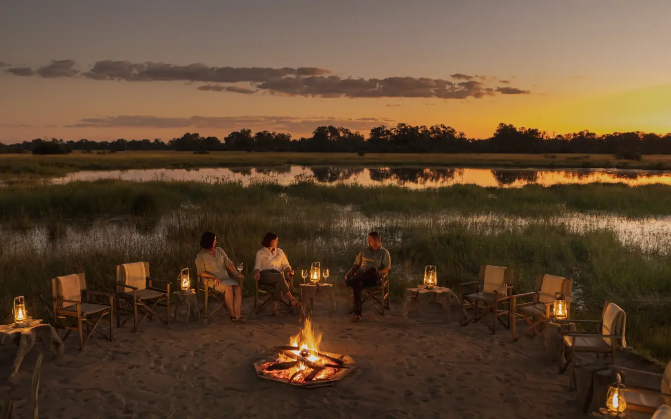 Guests gathered around evening campfire by wetlands at sunset, Sable Alley, The Okavango Delta, Botswana
