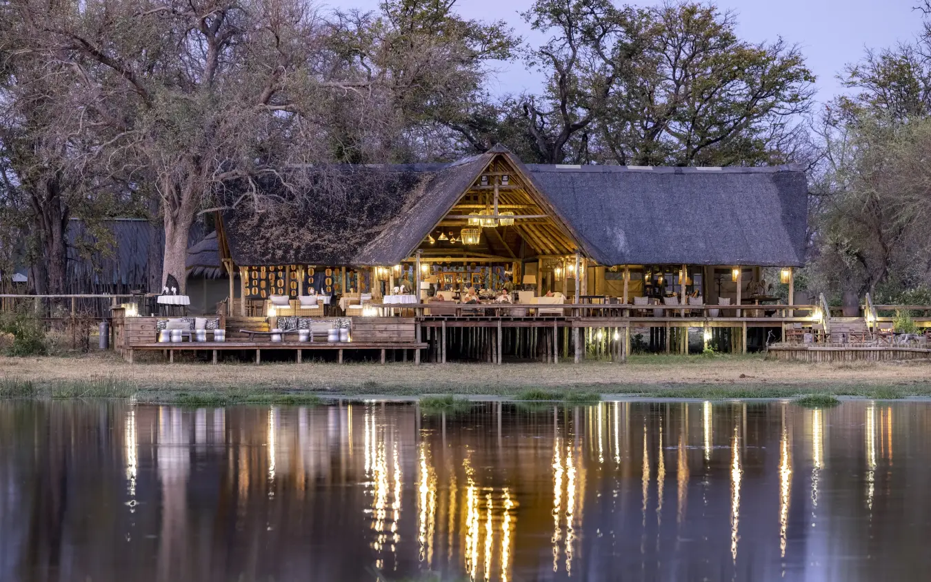 Thatched lodge buildings lit at dusk reflected in water, Sable Alley, The Okavango Delta, Botswana