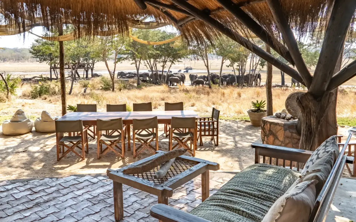 Outdoor dining area with fire pit under acacia trees overlooking elephants grazing, Sable Valley Lodge, Hwange National Park, Zimbabwe
