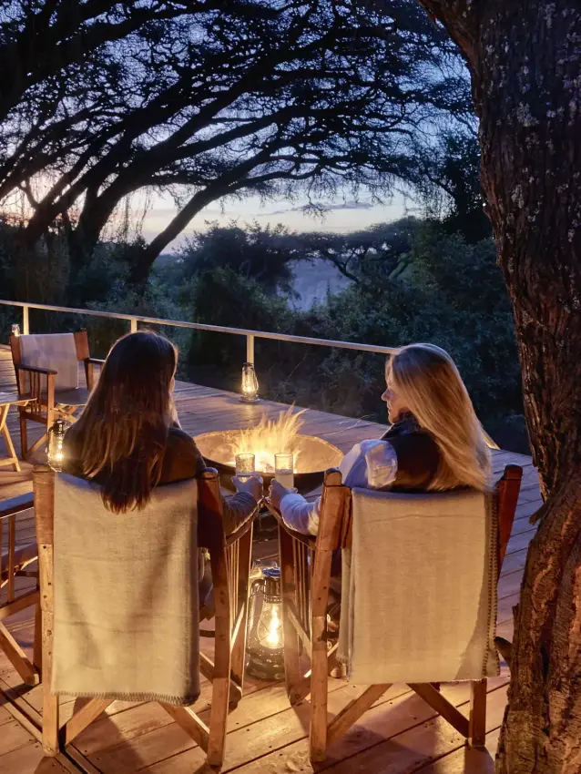 Two guests seated by fire pit on wooden deck at dusk, Sanctuary Crater Camp, Ngorongoro Crater, Tanzania