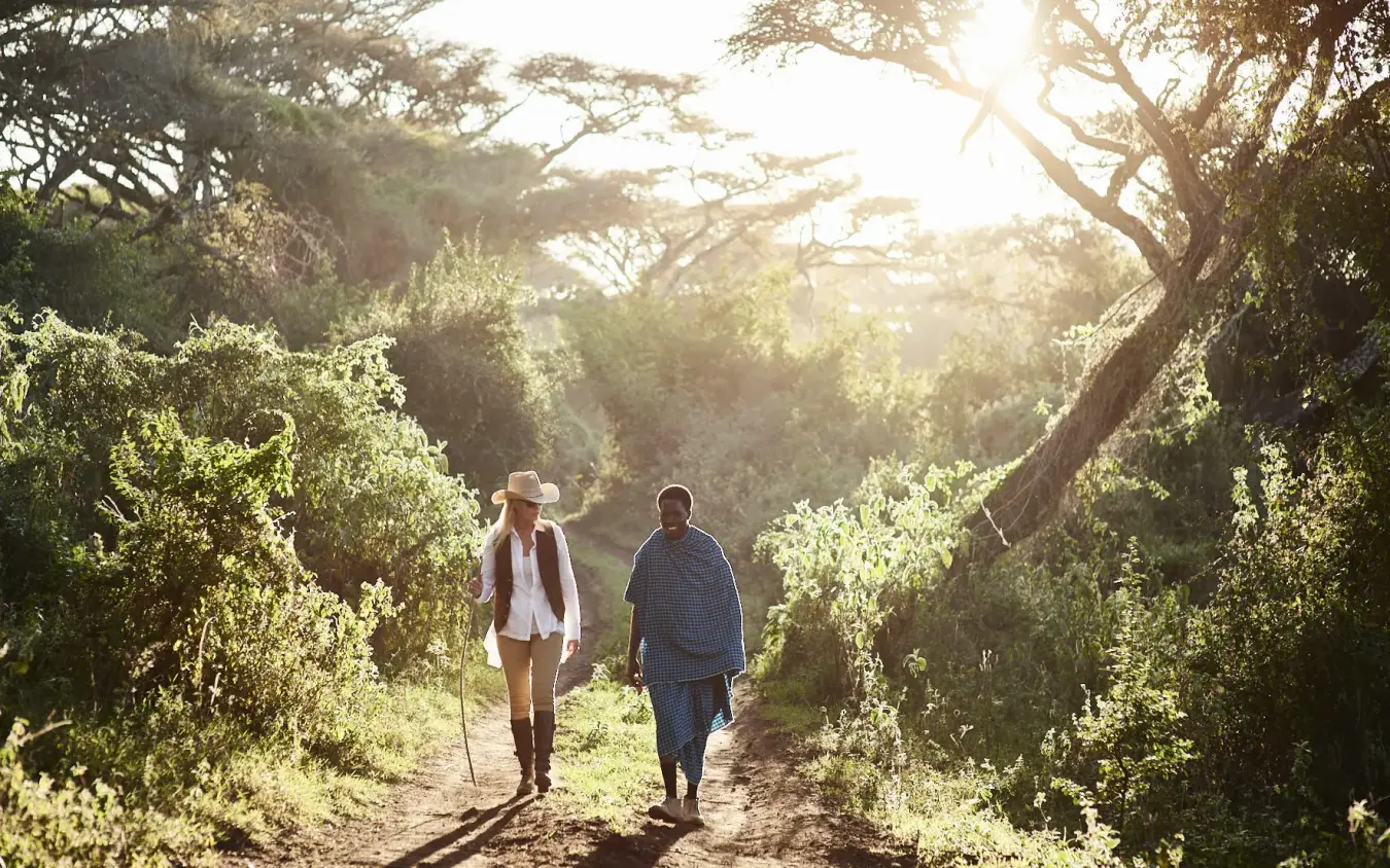Two hikers on tree-lined path at sunrise with dappled light, Sanctuary Crater Camp, Ngorongoro Crater, Tanzania