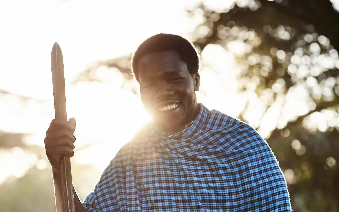 Smiling man in blue checkered cloth holding wooden spear, Sanctuary Crater Camp, Ngorongoro Crater, Tanzania