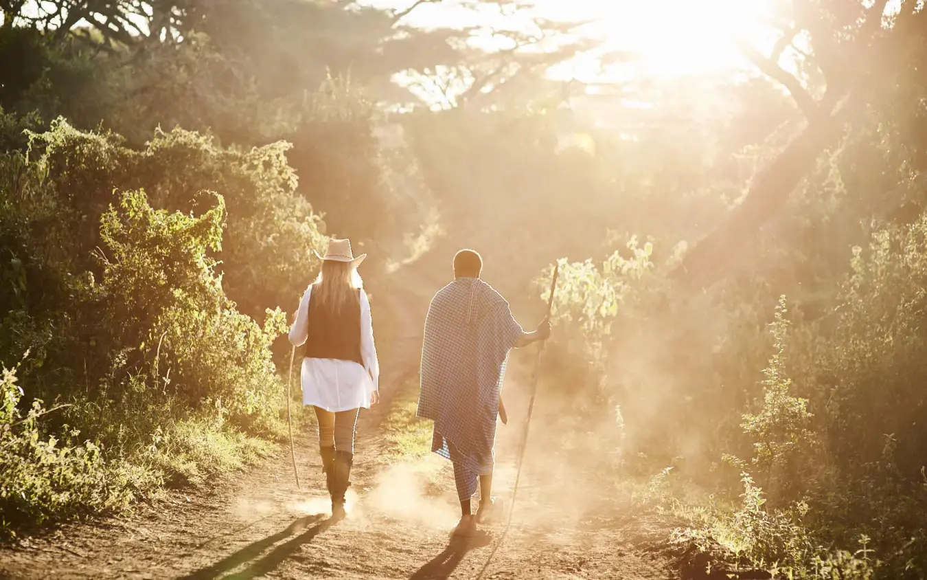 Two guests walking on dusty path through vegetation at sunrise, Sanctuary Crater Camp, Ngorongoro Crater, Tanzania