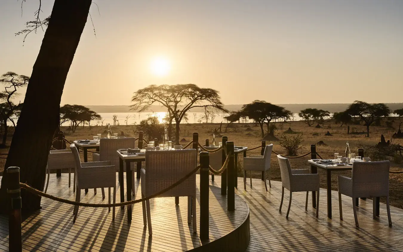 Outdoor dining deck with woven chairs overlooking acacia trees and waterhole at sunset, Swala, Tarangire National Park, Tanzania