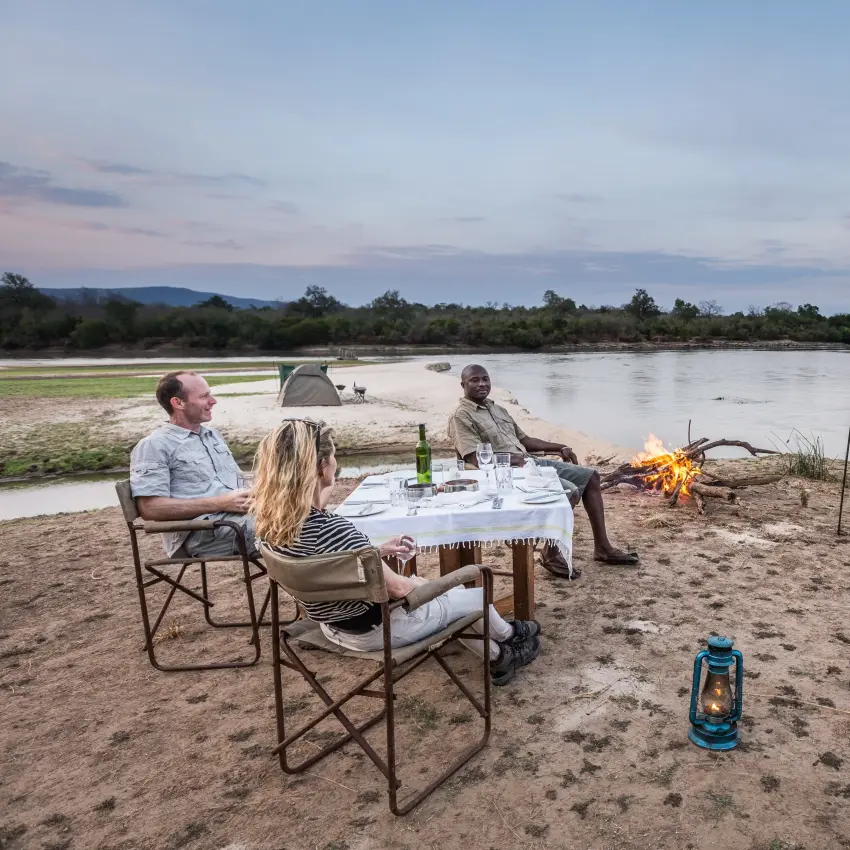 Guests dining at table by campfire on riverbank at dusk, Sand Rivers, Nyerere National Park, Tanzania
