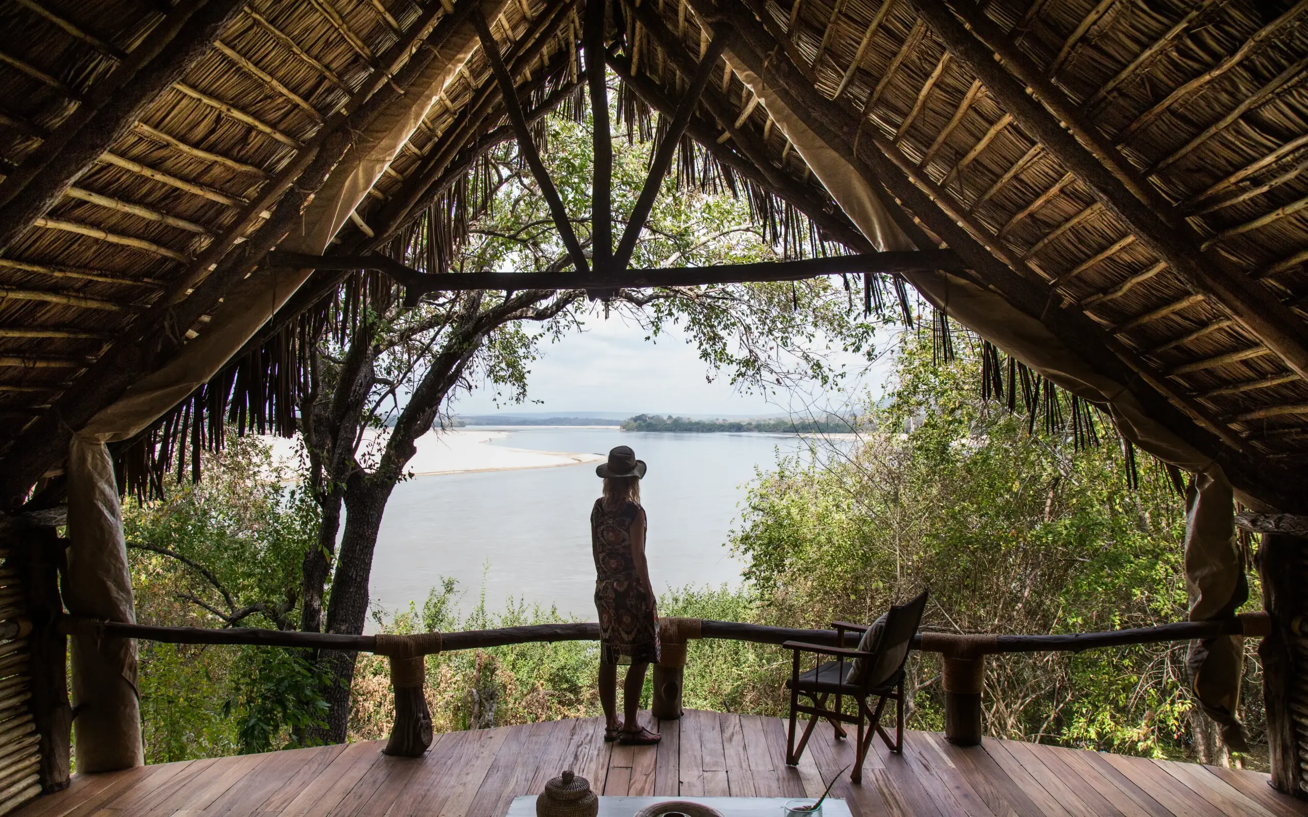 Person standing on wooden deck overlooking river from thatched pavilion, Sand Rivers, Nyerere National Park, Tanzania