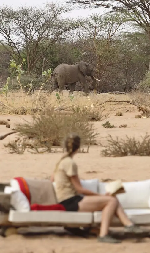 Woman relaxing in boat on sandy riverbank with elephant in background, Saruni Rhino, Northern Kenya, Kenya