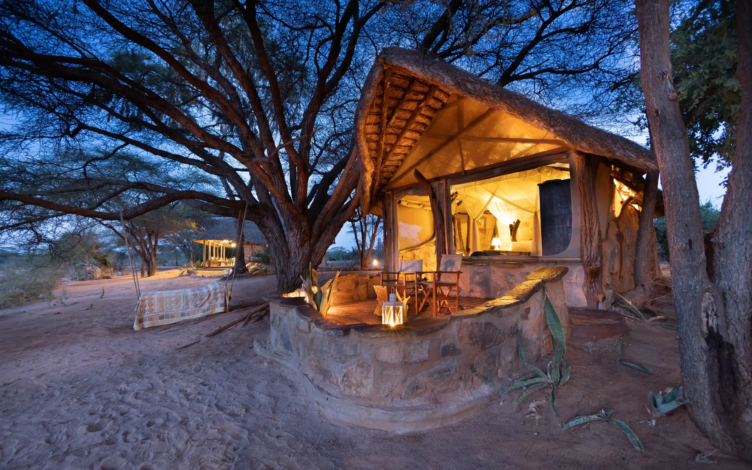Illuminated safari tent under ancient trees at dusk with sandy clearing, Saruni Rhino, Northern Kenya, Kenya