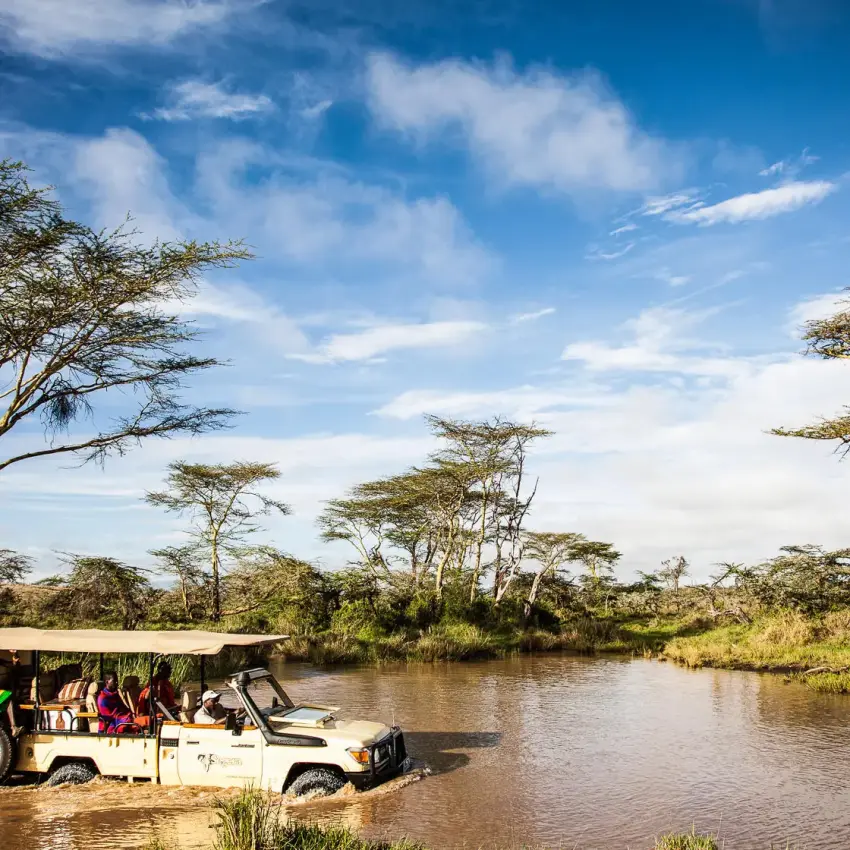 Safari vehicle crossing river with acacia trees and wildlife, Segera, Laikipia Highlands, Kenya