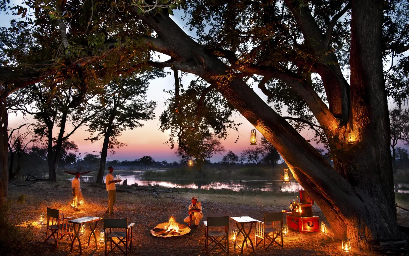Guests gather around campfire at sunset by river, Selinda Explorers Camp, Selinda Reserve, Botswana