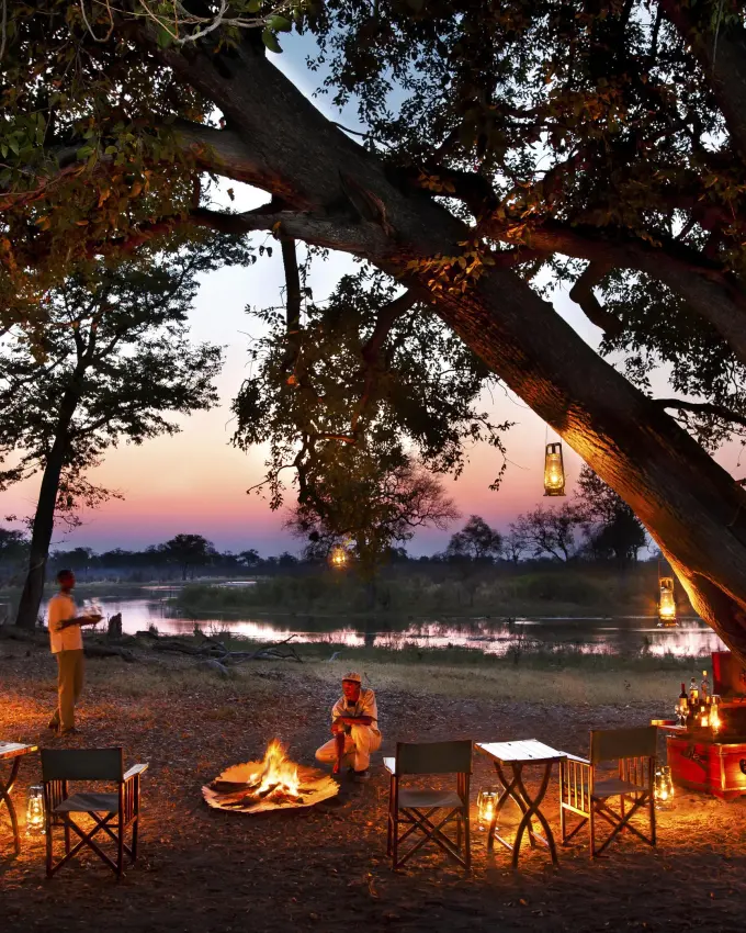 Guests gather around campfire at sunset by river, Selinda Reserve, Botswana