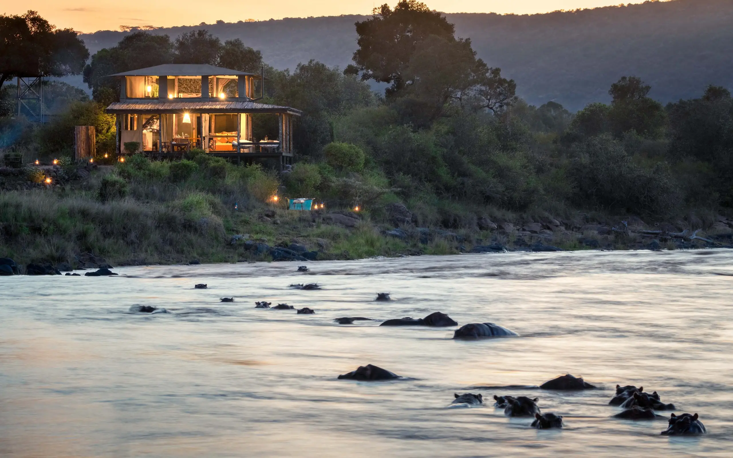 Riverside lodge at dusk with hippos in water, Kimya Kimya Camp, Maasai Mara, Kenya