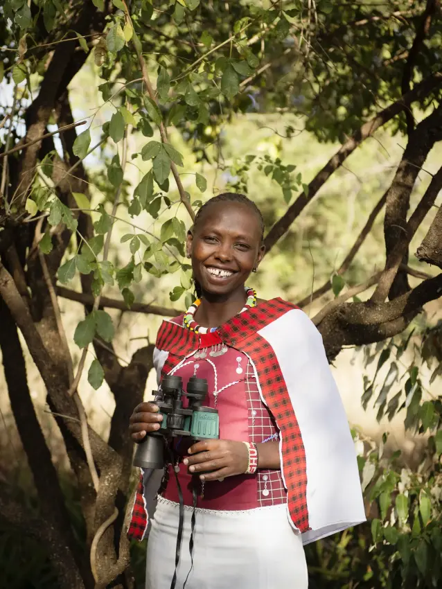 Young woman holding binoculars under tree shade, Serian “The Original”, Maasai Mara, Kenya