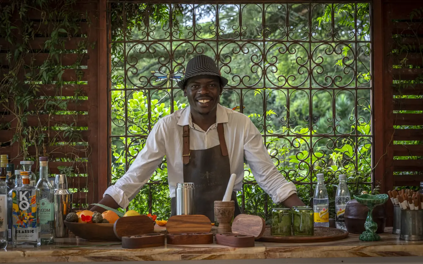Bartender smiling behind wooden counter with ornate window, Sheba’s Secret Garden, Nairobi, Kenya