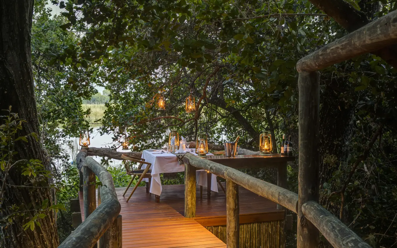 Wooden deck dining setup under trees at dusk, Shinde, The Okavango Delta, Botswana