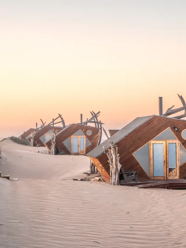 Wooden shipwreck-style structures at sunset across desert sand, Shipwreck Lodge, Skeleton Coast, Namibia