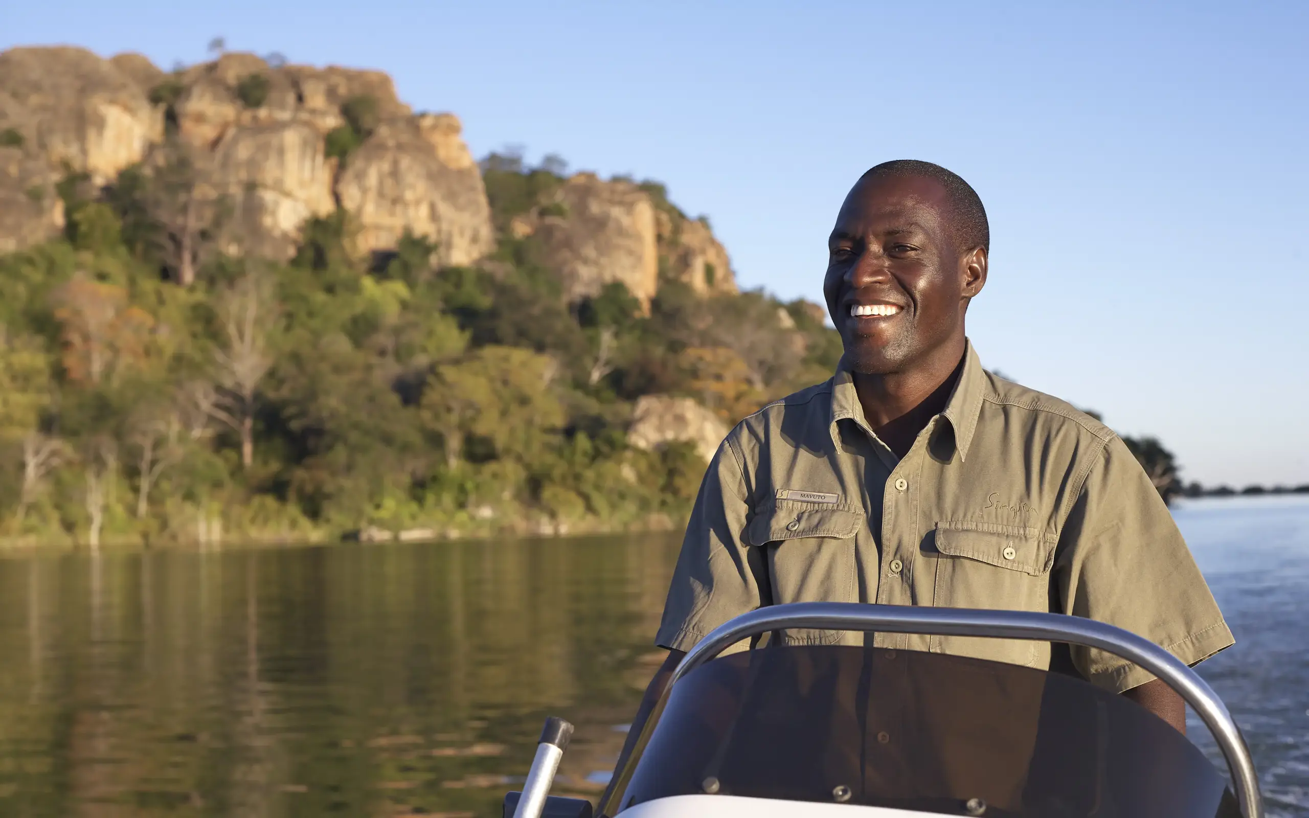 Man at boat helm on river with forested cliff backdrop, Zimbabwe