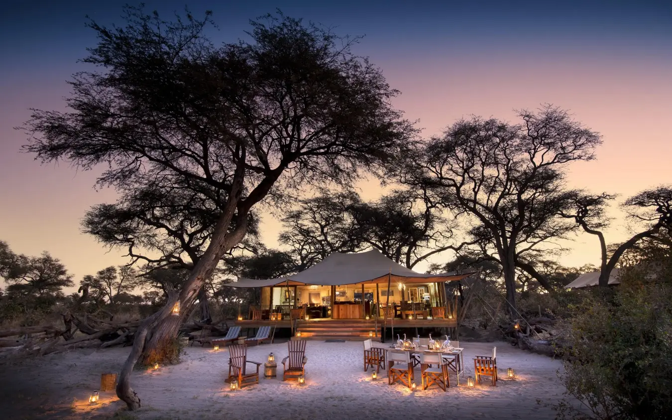 Canvas tent lodge at dusk with acacia trees and candlelit seating, Somalisa Expeditions, Hwange National Park, Zimbabwe