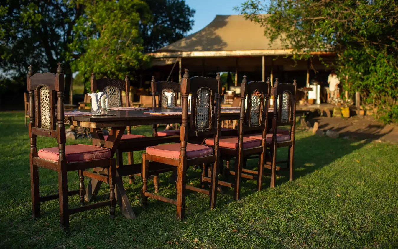 Outdoor dining setup with wooden chairs and table on grass, Speke’s Camp, Maasai Mara, Kenya
