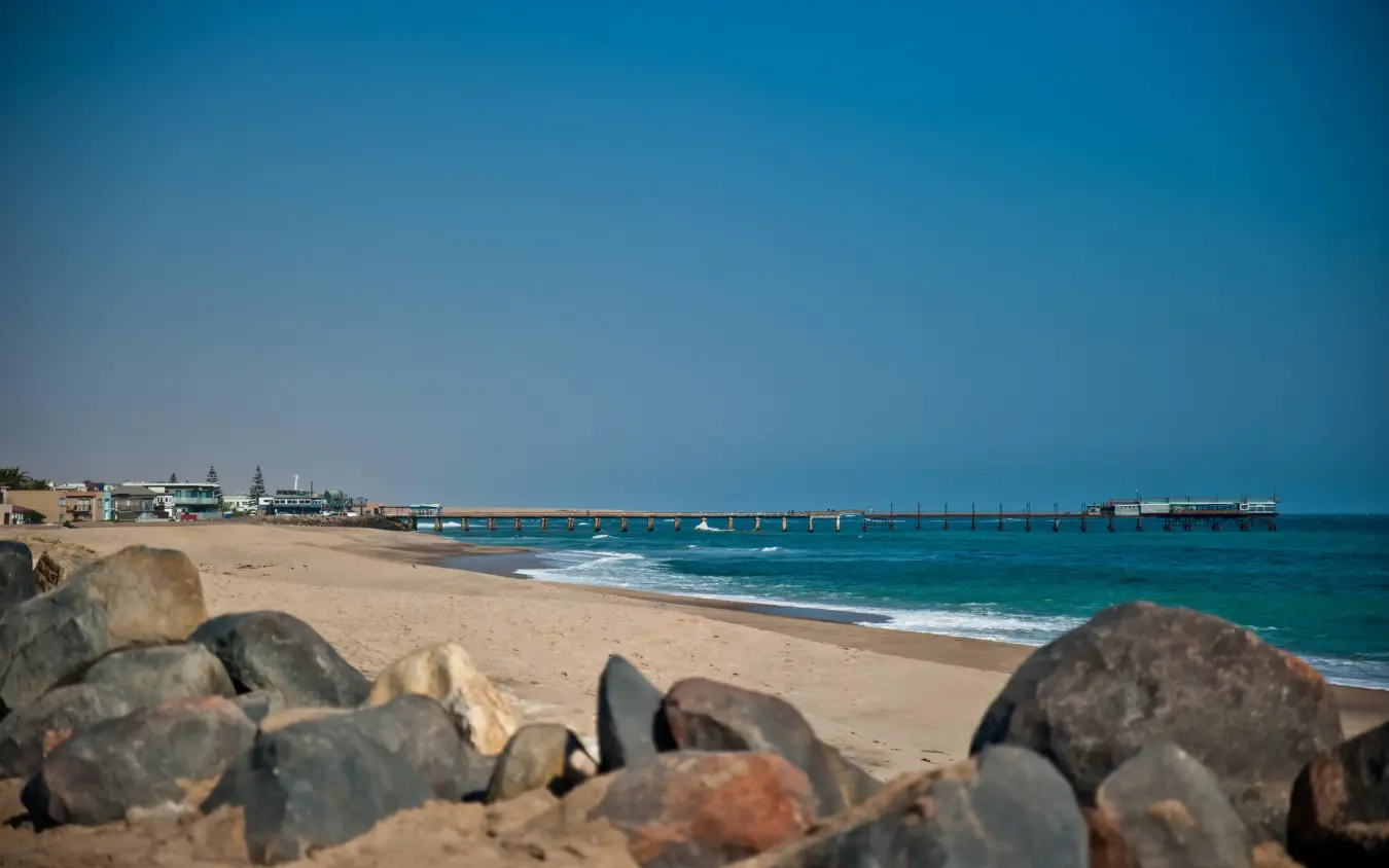 Sandy beach with rocks and long pier extending into turquoise ocean, The Delight Swakopmund, Swakopmund, Namibia