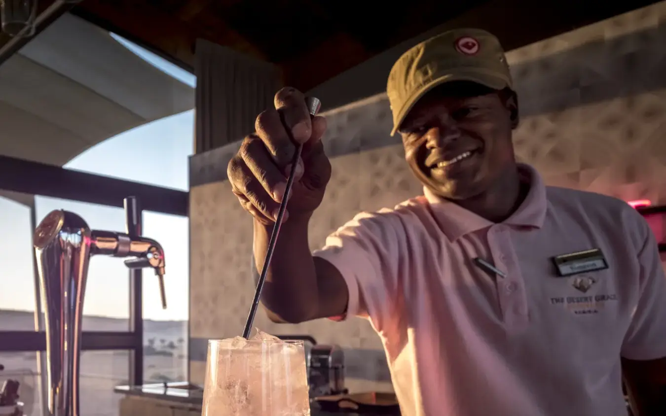 Bartender mixing cocktail at outdoor counter with desert landscape view, The Desert Grace, Sossusvlei, Namibia