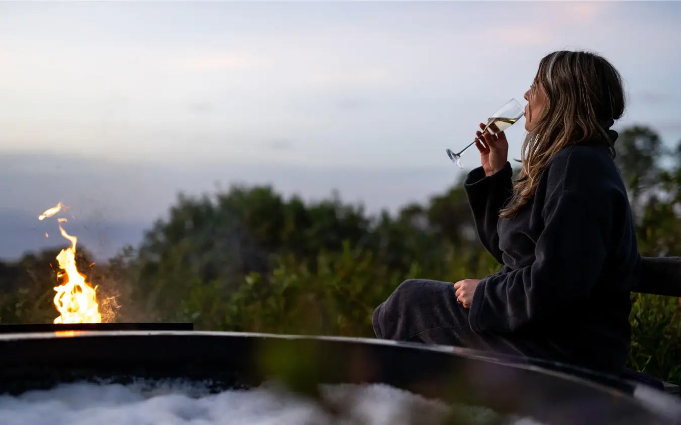Woman enjoying wine by fire overlooking misty landscape, The Highlands, Ngorongoro Crater, Tanzania