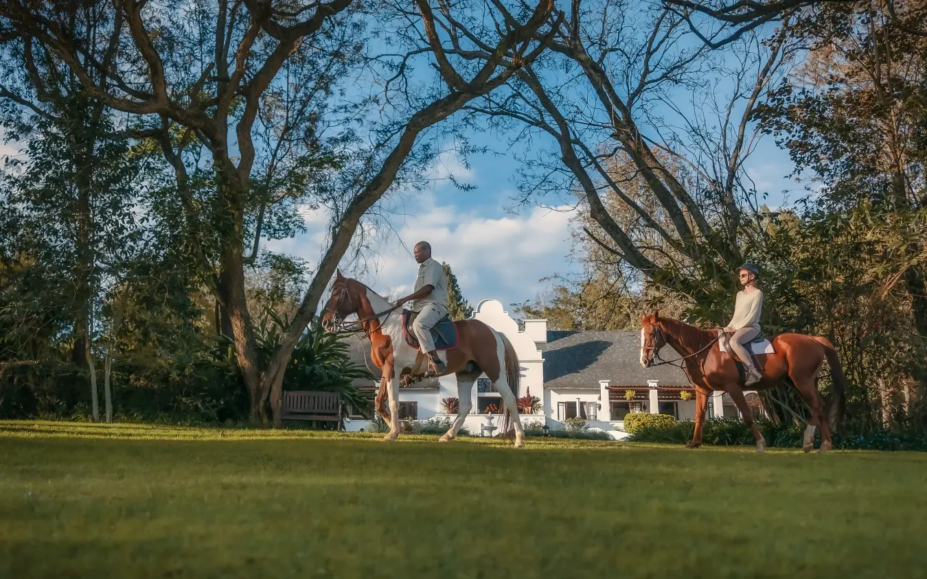 Horseback riders on manicured lawn with colonial-style mansion and tree canopy, The Manor at Ngorongoro, Ngorongoro Crater, Tanzania