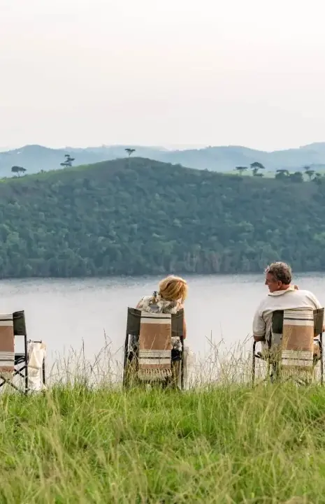 Guests in camping chairs overlooking river and forested banks, The River Station, Queen Elizabeth NP, Uganda