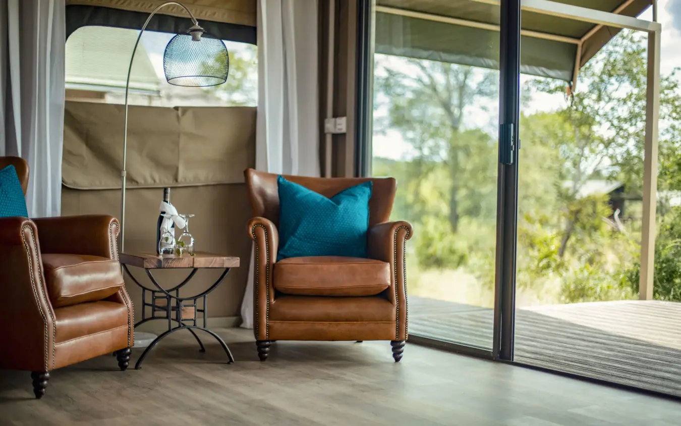 Leather armchairs with blue cushions overlooking trees through glass doors, The Wallow Lodge, Hwange National Park, Zimbabwe