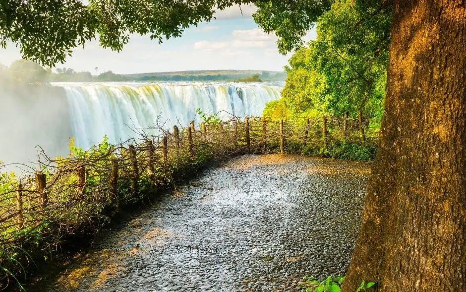 Waterfall viewed from tree-lined pathway with wooden fence, The Wallow Lodge, Hwange National Park, Zimbabwe