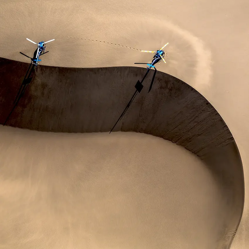 Aerial view of two helicopters hovering above desert sand dune with shadow