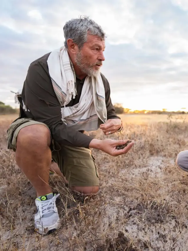 Two men crouching in dry grassland having conversation, Usangu Expedition Camp, Ruaha National Park, Tanzania