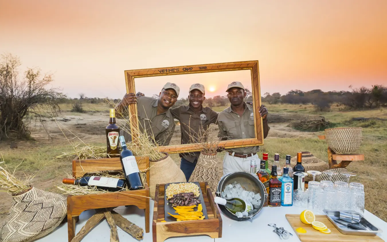 Three men holding wooden frame at outdoor dining setup during sunset, Verney’s Camp, Hwange National Park, Zimbabwe