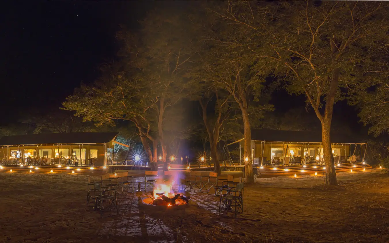 Nighttime safari camp with firepit, seated guests, and illuminated lodge structures, Verney’s Camp, Hwange National Park, Zimbabwe