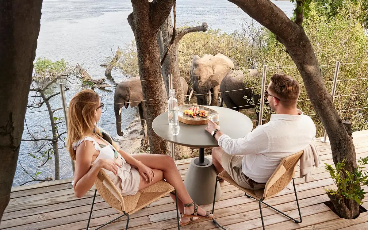 Couple dining on riverside deck with elephants nearby, Island Treehouse Suites, Victoria Falls, Zimbabwe