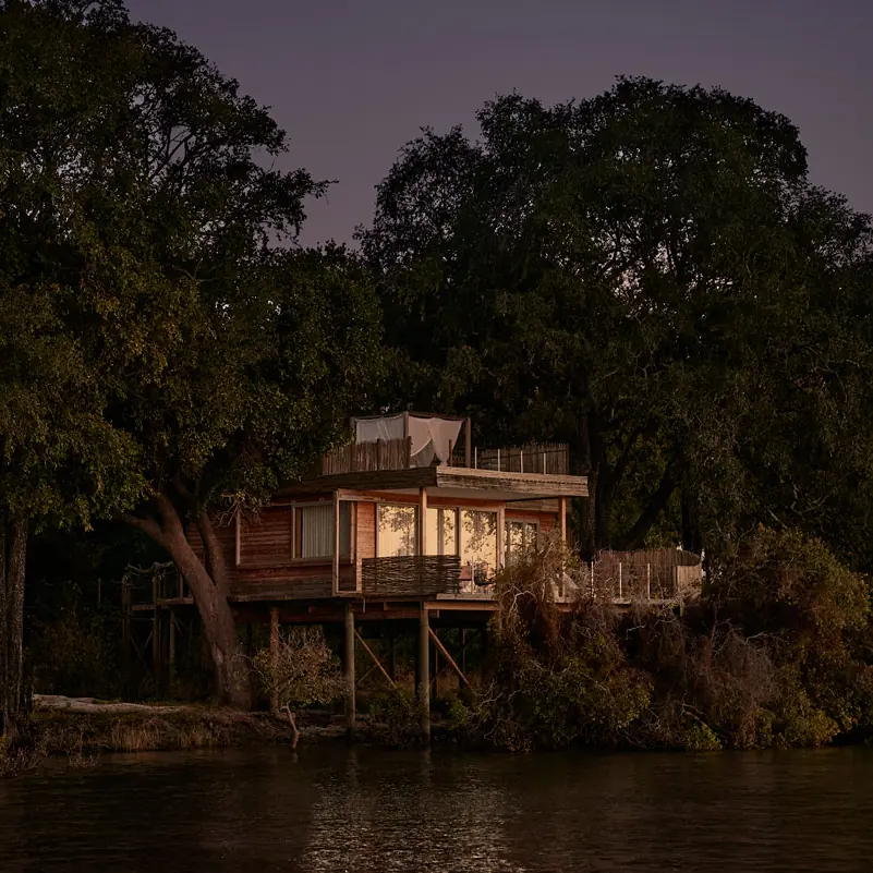 Wooden lodge structure illuminated at dusk among riverside trees, Island Treehouse Suites, Victoria Falls, Zimbabwe