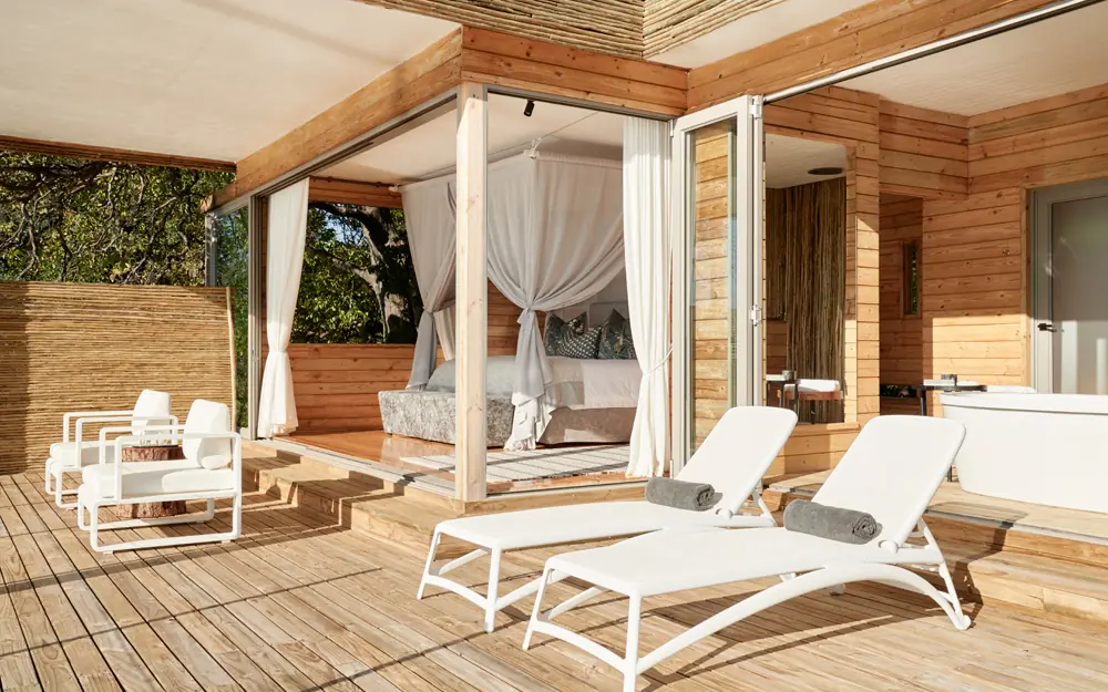 Wooden lodge bedroom with four-poster bed and deck lounge chairs, Island Treehouse Suites, Victoria Falls, Zimbabwe