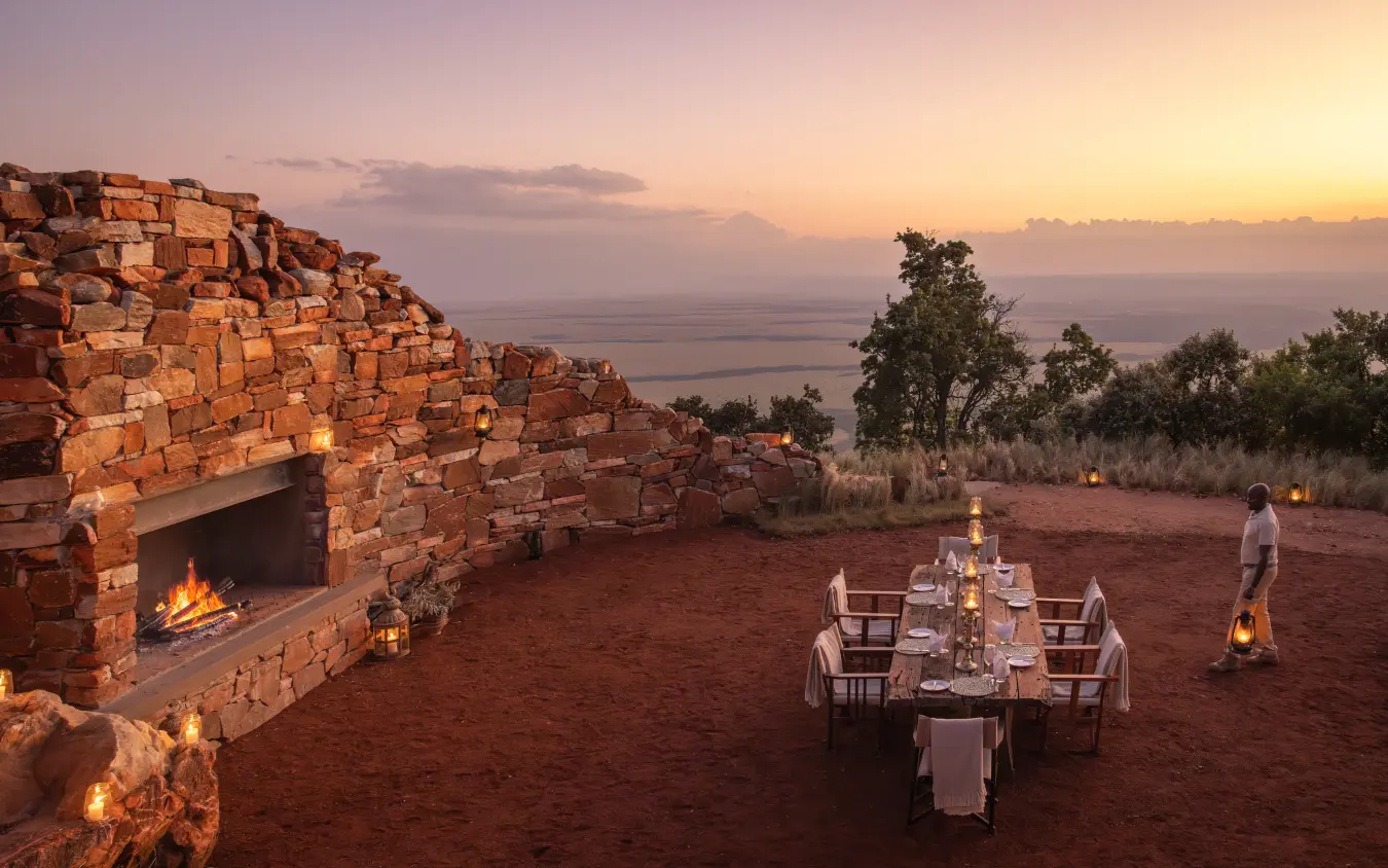 Outdoor dining setup at sunset with fireplace and water view, Wild Hill, Maasai Mara, Kenya
