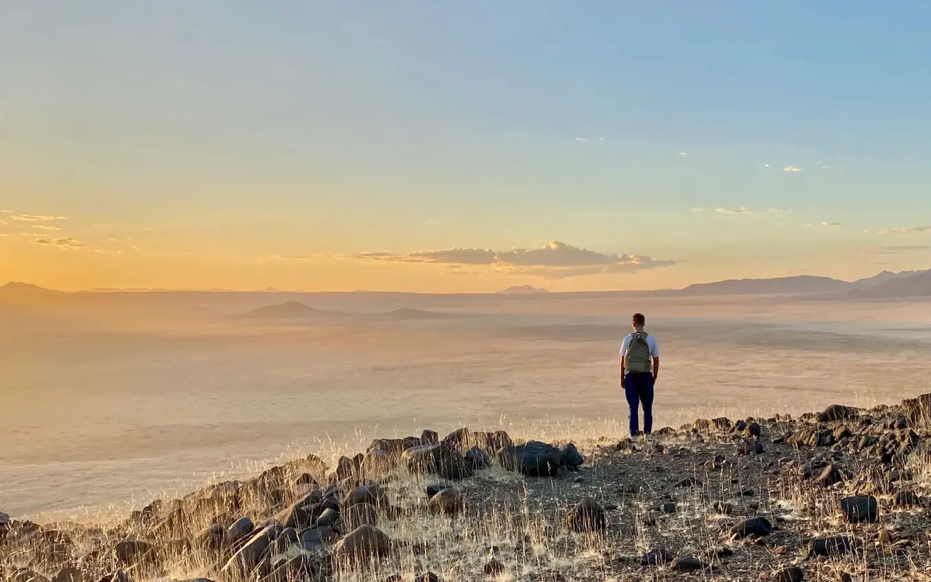 Hiker standing on rocky outcrop overlooking vast desert landscape, Wolwedans Boulders Camp, NamibRand Private Reserve, Namibia