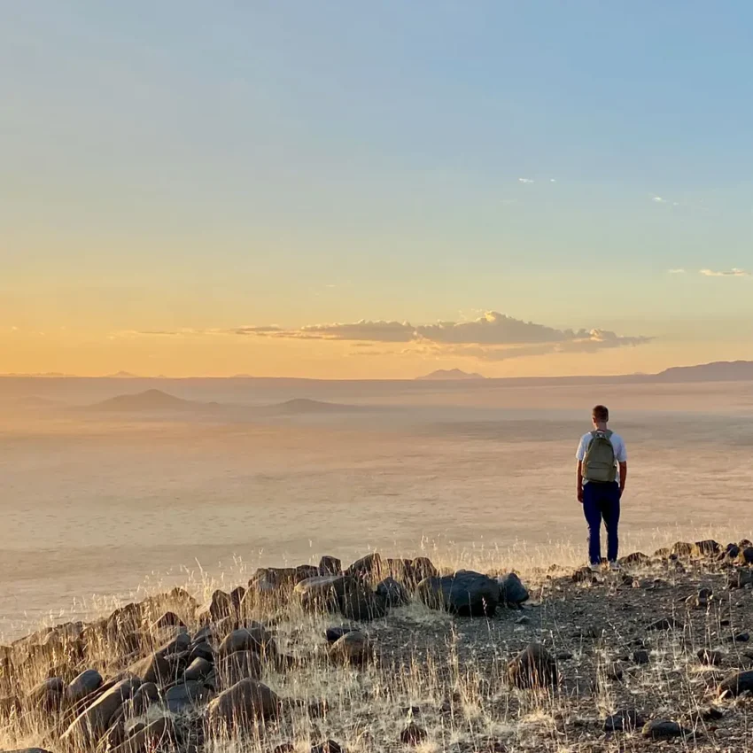 Hiker standing on rocky outcrop overlooking vast desert landscape, Wolwedans Boulders Camp, NamibRand Private Reserve, Namibia