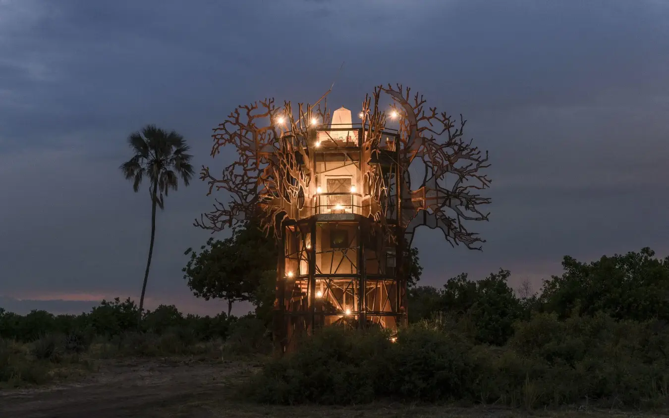 Illuminated treehouse structure at dusk with palm tree, Xigera Lodge, The Okavango Delta, Botswana