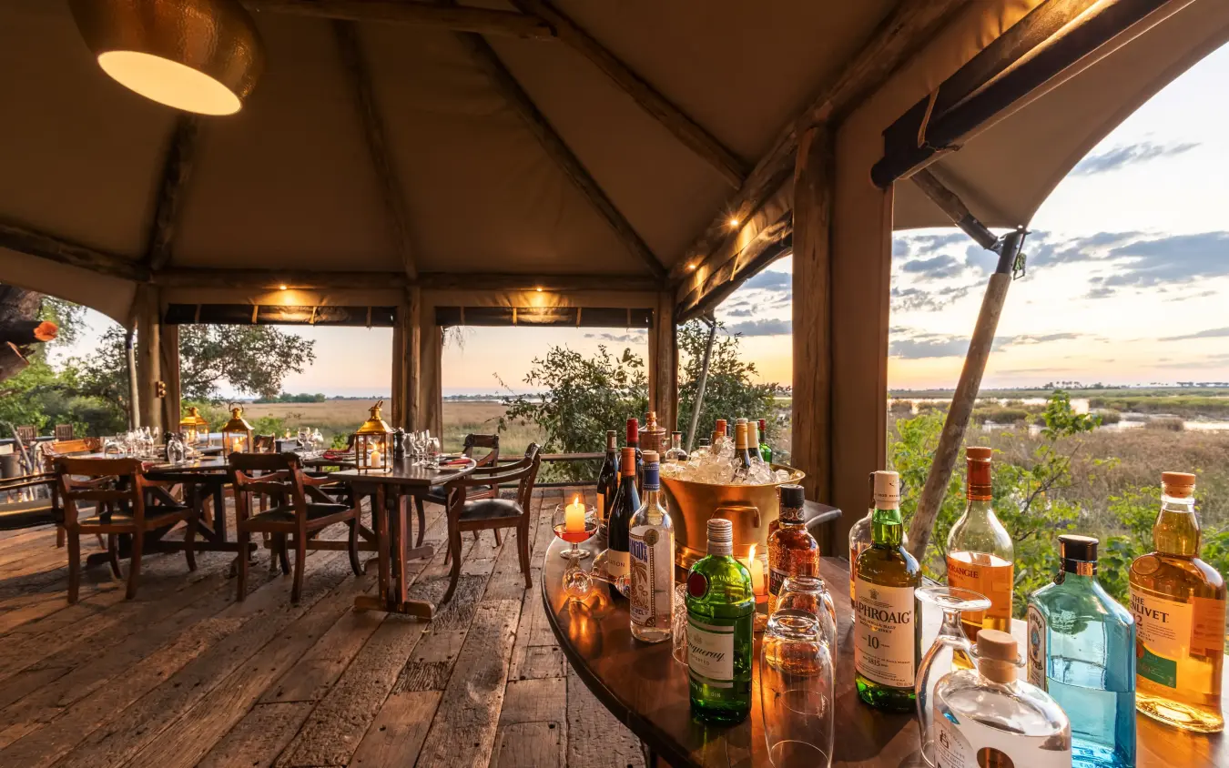 Elevated dining deck with bottles and sunset landscape view, Zarafa Camp, Selinda Reserve, Botswana