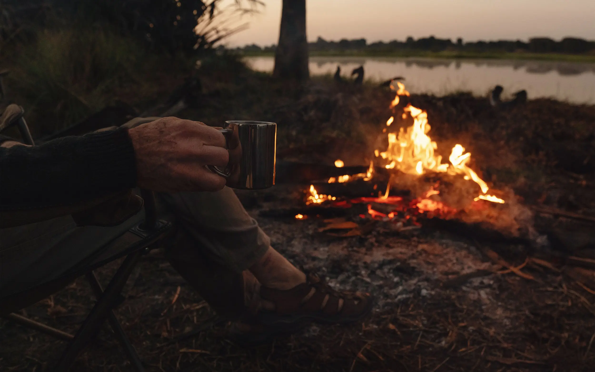 Person holding metal cup by campfire overlooking river at dusk, Charlie Ugalla, Ugalla River National Park, Tanzania