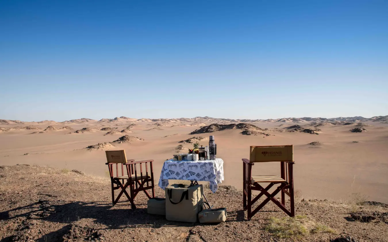Dining table set up in desert landscape with two chairs overlooking dunes, Hoanib Elephant Camp, Skeleton Coast, Namibia