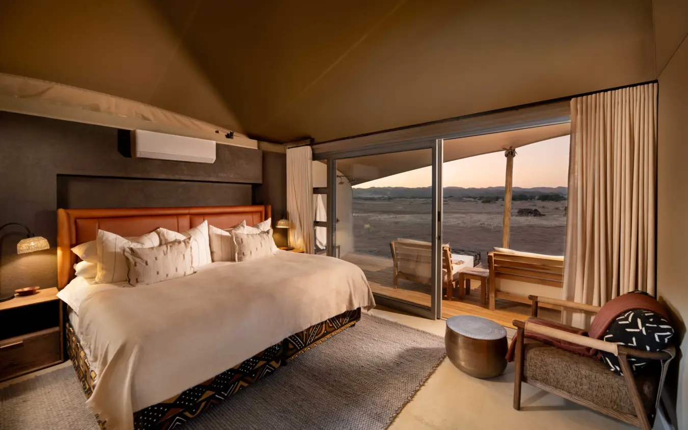 Luxury bedroom suite with panoramic windows overlooking vast savanna landscape at dusk, Hoanib Elephant Camp, Skeleton Coast, Namibia