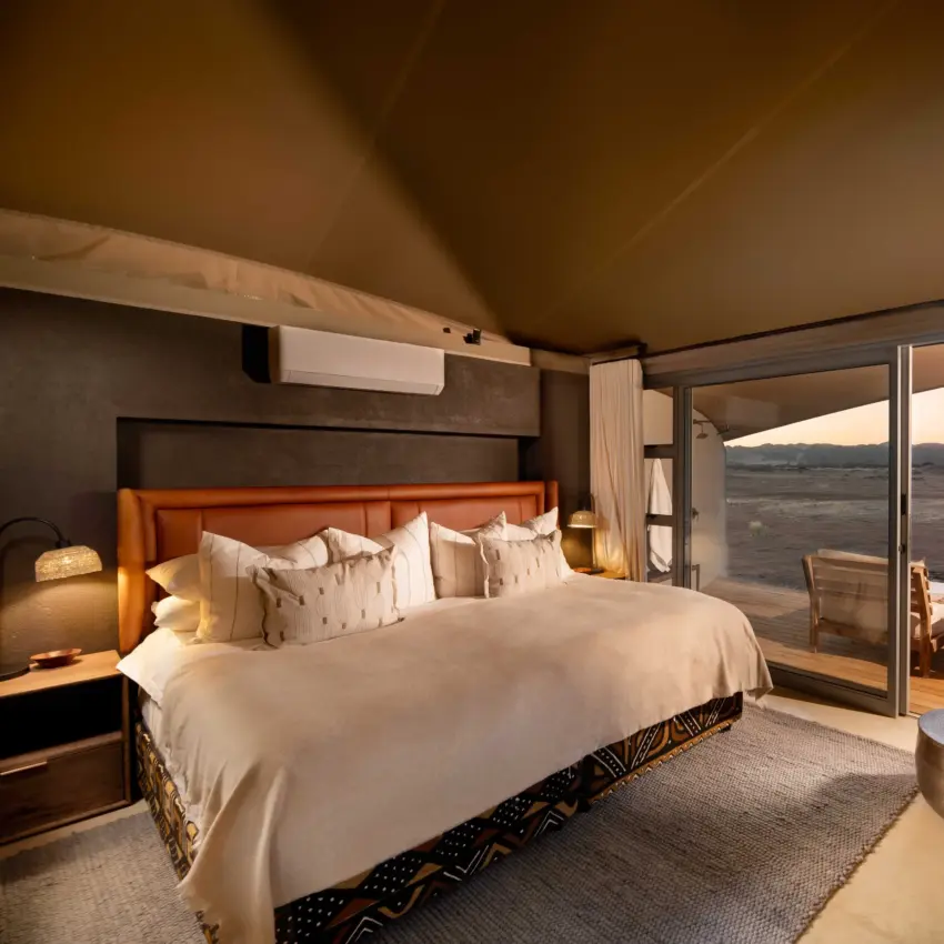 Luxury bedroom suite with panoramic windows overlooking vast savanna landscape at dusk, Hoanib Elephant Camp, Skeleton Coast, Namibia