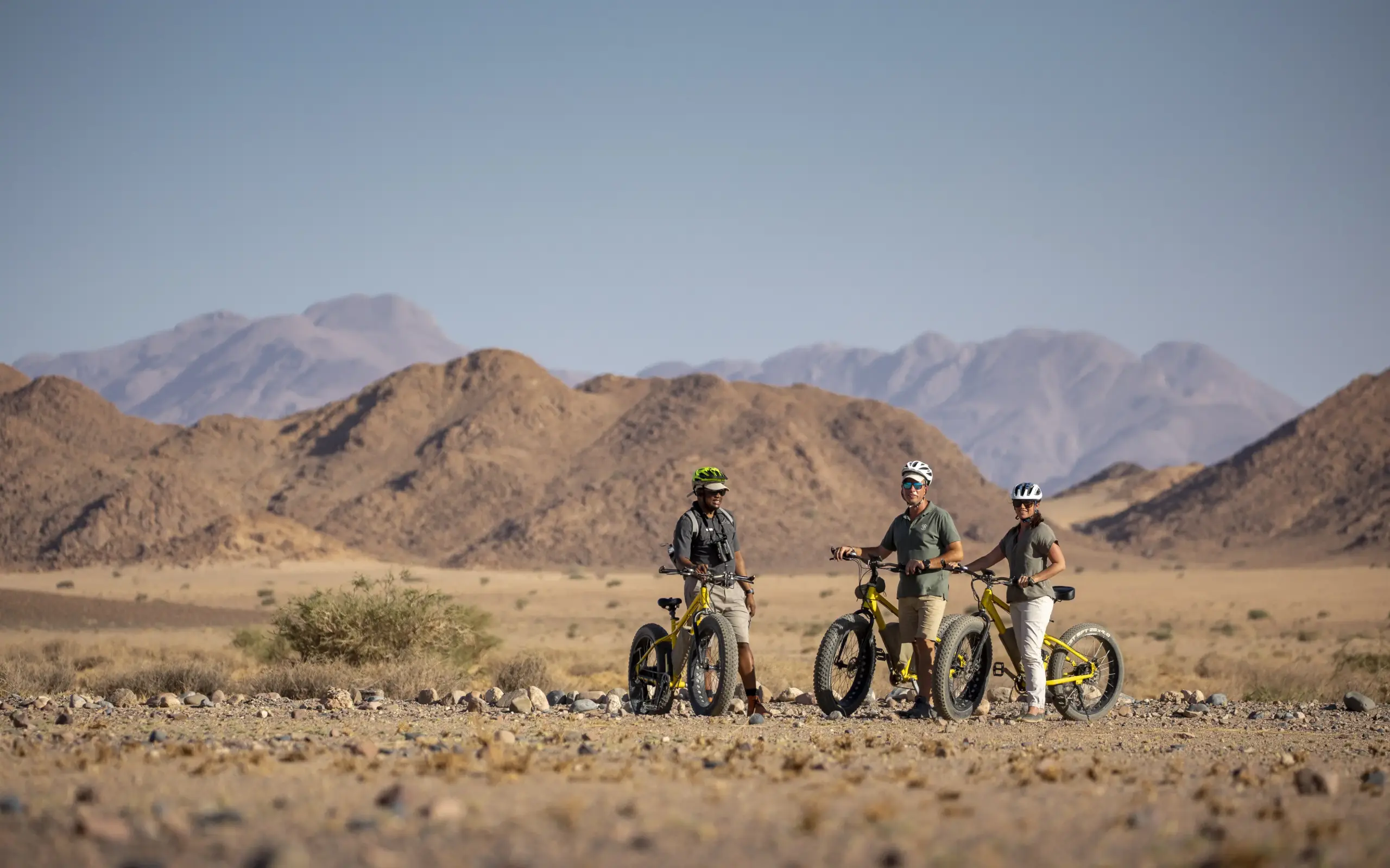 Mountain bikers with guide on desert plains beneath rocky peaks, Little Kulala, Sossusvlei, Namibia