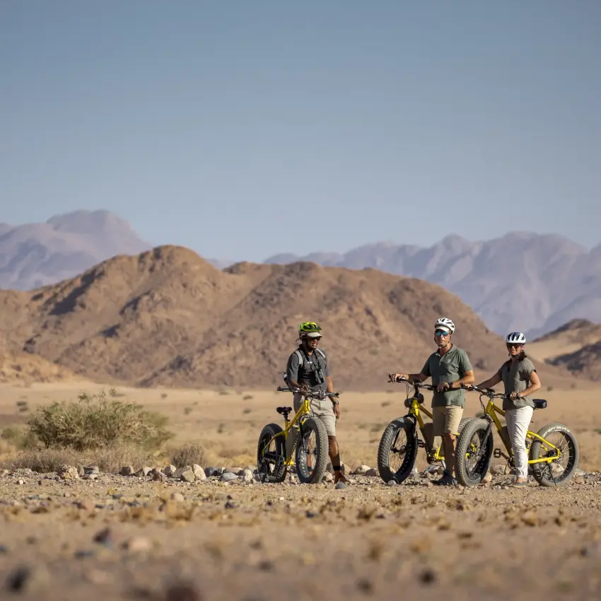 Mountain bikers with guide on desert plains beneath rocky peaks, Little Kulala, Sossusvlei, Namibia