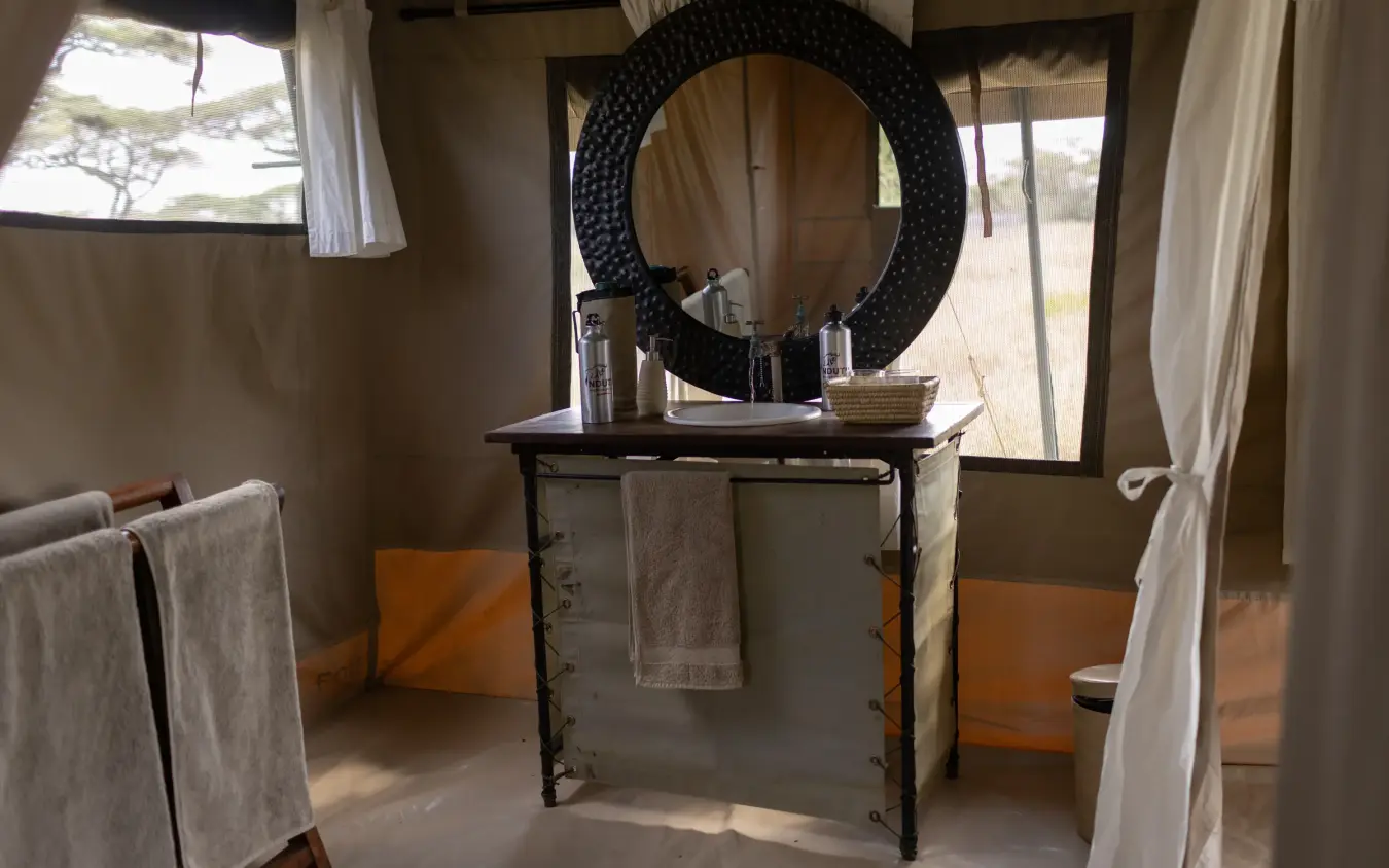 Luxury safari tent bathroom with round mirror and savanna views through windows, Ndutu Wilderness Camp, Serengeti National Park, Tanzania